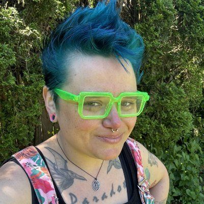 Sassafras, a white genderqueer person with bright blue teal hair lime green translucent square glasses and several tattoos visible including birds on either side of their collarbones